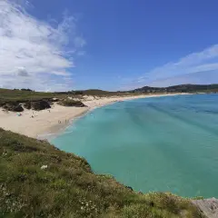 Vista panorámica de la playa de Santa Comba, con arena dorada, aguas turquesas y colinas verdes. Algunas personas disfrutan del sol y el mar.