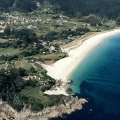 Vista aérea de la playa Area Brava, con arena blanca, aguas turquesas y un pueblo costero rodeado de vegetación y campos verdes.