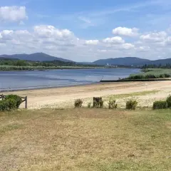 La playa fluvial Vilarello, con arena dorada y aguas tranquilas, se extiende ante colinas verdes bajo un cielo azul con nubes blancas.