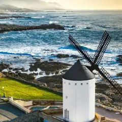 Un muiño do vento blanco con aspas negras se alza junto a un mar azul y rocoso. El paisaje costero es agreste y hermoso.