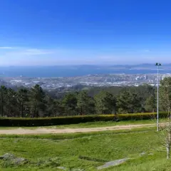 Vista panorámica desde el monte dos pozos, con un camino de tierra y césped en primer plano, árboles y un faro, y una ciudad y el mar al fondo.