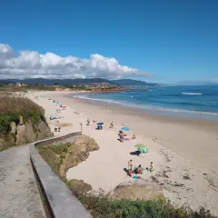 Un paseo de piedra bordea la **playa llas**, donde gente descansa bajo sombrillas y el mar azul invita a un baño.