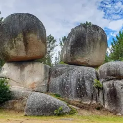 Gigantescas rocas apiladas, como penas de rodas, desafían la gravedad en un paisaje boscoso bajo un cielo azul con nubes.