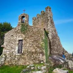 Antigua ermita de piedra con campanario y cruz, rodeada de vegetación. Las **torres de oeste** se alzan imponentes contra el cielo azul.