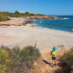 Niño con mochila azul desciende por sendero hacia la playa de rueta, con rocas y mar turquesa.