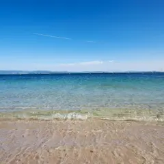 Aguas cristalinas en la playa milide, con veleros navegando en el horizonte bajo un cielo azul intenso.