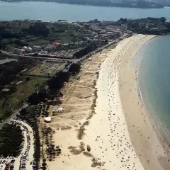 Vista aérea de una playa grande y concurrida, con arena dorada, mar azul y un pueblo costero al fondo.