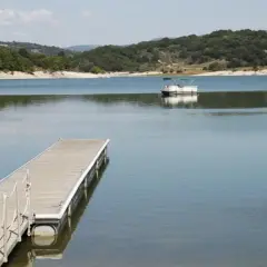 Un pontón blanco navega en el tranquilo embalse de Prada, con un muelle de madera en primer plano y colinas verdes al fondo.