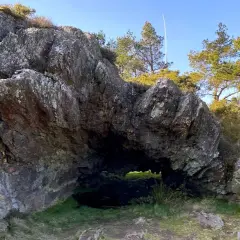 Entrada a la cova da serpe, una cueva natural entre rocas y vegetación bajo un cielo azul.