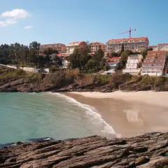 La **playa Caneliñas** se asoma entre rocas y vegetación, con edificios residenciales en la colina y el mar turquesa.