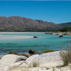 Aguas turquesas y arena blanca bordean la costa, con el imponente monte Pindo como telón de fondo. Rocas cubiertas de musgo salpican la orilla.