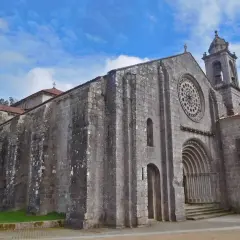 El monasterio de Armenteira, con su imponente fachada de piedra y su rosetón, se alza bajo un cielo azul con nubes.