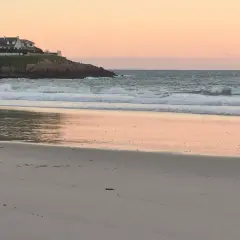 Ocaso en la playa areoura. Olas rompen suavemente en la arena dorada, reflejando el cielo rosado. Casas blancas coronan el acantilado.