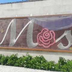 Letras de encaje blanco y rosa forman "Mos" en un letrero en el Pazo de Mos, con herramientas de encaje a la izquierda.