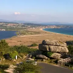 Vista panorámica desde el mirador de Siradella. Rocas gigantescas y un puente de madera en primer plano, con un paisaje de costa, mar azul y un pueblo al fondo.