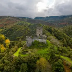 El **castillo de Doiras** se alza majestuoso sobre una colina verde, rodeado de montañas boscosas y un cielo nublado.
