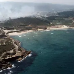 Vista aérea de la playa de Sabón, con una chimenea industrial y humo en el fondo, y el mar azul turquesa.