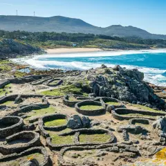 Ruinas circulares de piedra del castro de Baroña, con el mar azul y una playa de arena al fondo.