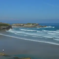 La **playa Os Castros** se extiende bajo un cielo azul, con olas rompiendo en la arena dorada. Una casa moderna corona el acantilado.