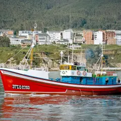 El barco museo boniteiro Reina del Carmen navega con su casco rojo y azul, reflejándose en el agua.