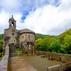 Antiguo monasterio de San Juan de Caaveiro, con su torre campanario y ábside románico, rodeado de exuberante vegetación verde.