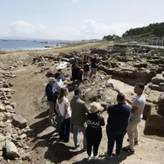 Grupo de personas observando ruinas arqueológicas en la costa. Se aprecia el mar y el **adro vello** del yacimiento.