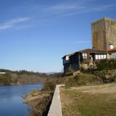 La torre de Lapela se alza junto a un río, con casas antiguas a su lado y un cielo azul despejado.