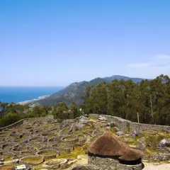 Ruinas circulares de piedra en el monte Santa Tecla, con el mar y montañas al fondo.