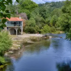 Disfrutando del sol y el agua en la playa fluvial Mondariz. Un edificio con arcos y tejado rojo se alza junto al río, donde unas personas se refrescan.
