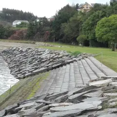 Escaleras de piedra y césped verde junto al agua en la playa de los bloques Ribadeo, con casas y árboles al fondo.