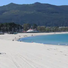 Amplia playa de arena blanca en la playa de Samil, con aguas turquesas y una montaña verde al fondo.