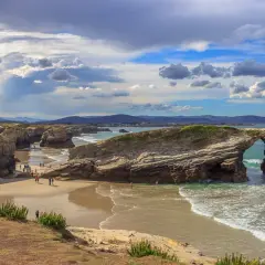Espectacular arco natural en la playa de Esteiro, Ribadeo. Olas rompen en la arena dorada bajo un cielo nublado.