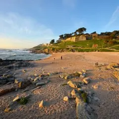 Atardecer en la playa concheira de Baiona. Olas rompen en la costa rocosa, mientras una fortaleza histórica corona la colina verde.