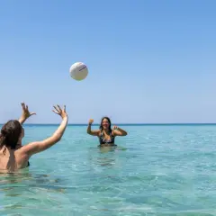Dos amigas juegan voleibol en el mar turquesa de la playa América, bajo un cielo azul despejado.
