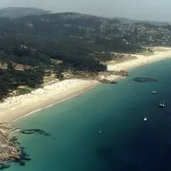 Vista aérea de la playa de Nerga, con arena dorada, aguas turquesas y barcos navegando. El paisaje se completa con pinos y colinas verdes.