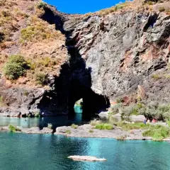 Un río de aguas turquesas fluye hacia un monte furado, una cueva natural en la roca, con vegetación y personas disfrutando del paisaje.