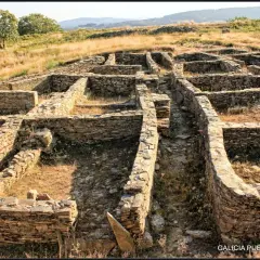 Ruinas de piedra del castro de Castromaior, con muros bajos y patios interiores, bajo un cielo azul y vegetación seca.