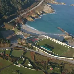 Vista aérea de un campo de fútbol junto a una playa a marosa, con el mar azul y rocas.