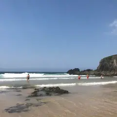 Gente disfrutando del mar en la playa de Frouxeira, con olas rompiendo y una gran roca verde al fondo bajo un cielo azul.