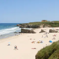 Gente disfrutando del sol y el mar en la playa Longara Barreiros, con formaciones rocosas y una casa amarilla al fondo.