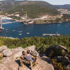 Pareja sentada en un mirador, contemplando el impresionante puente de Rande sobre el mar, con bateas de mejillones y un pueblo al fondo.