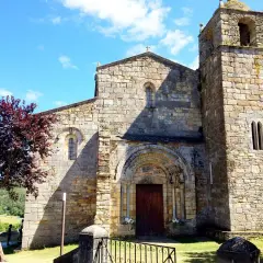 La catedral de Foz, con su torre románica y fachada de piedra, se alza bajo un cielo azul con nubes. Un árbol de hojas rojizas añade color.