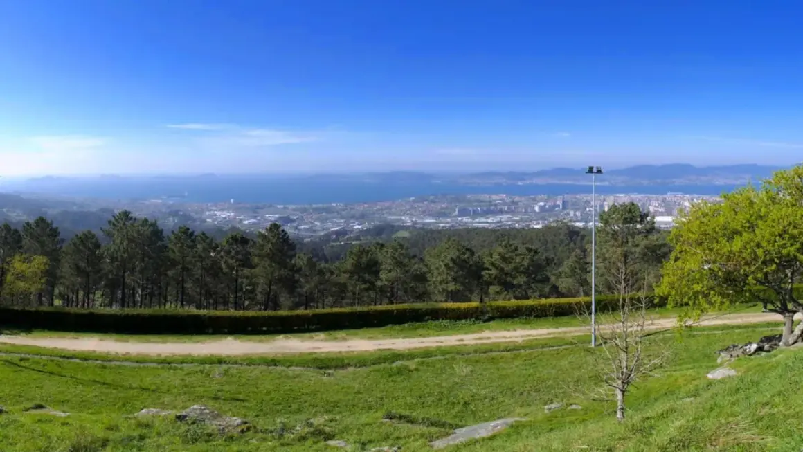 Vista panorámica desde el monte dos pozos, con un camino de tierra y césped en primer plano, árboles y un faro, y una ciudad y el mar al fondo.