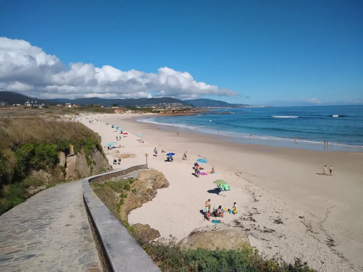 Un paseo de piedra bordea la **playa llas**, donde gente descansa bajo sombrillas y el mar azul invita a un baño.
