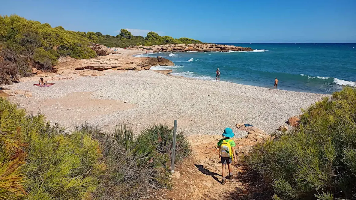 Niño con mochila azul desciende por sendero hacia la playa de rueta, con rocas y mar turquesa.