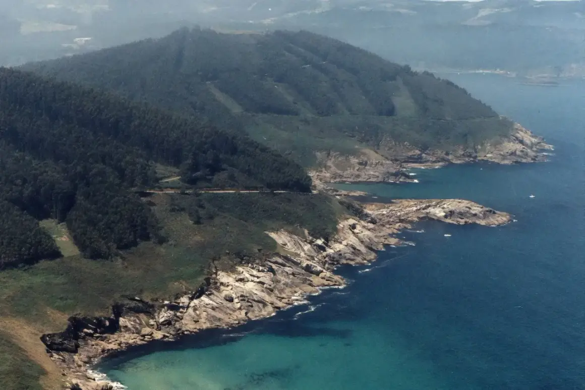 Costa rocosa y verde con el mar azul turquesa. Se ve una carretera serpenteando por la ladera. La playa Portonovo Viveiro es un paraíso natural.