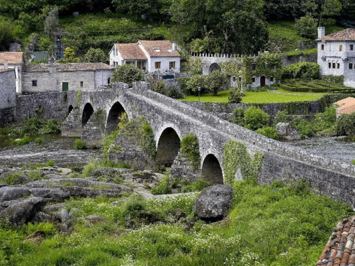 Un antiguo ponte carreira de piedra cruza un río, rodeado de casas rústicas y vegetación exuberante.