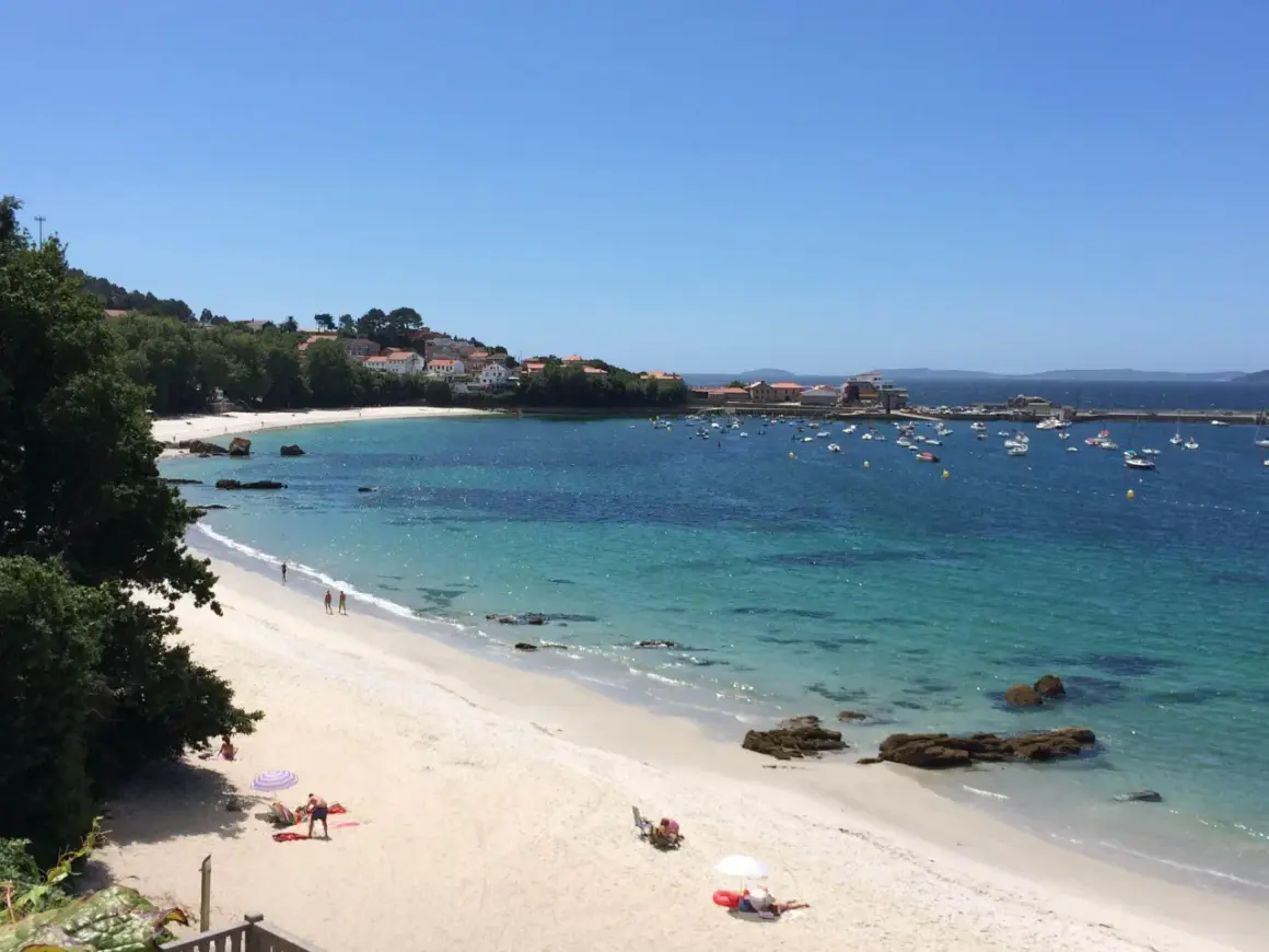 La **playa de Aguete** luce un día soleado con aguas cristalinas y barcos fondeados. Gente disfruta de la arena blanca y el mar azul.