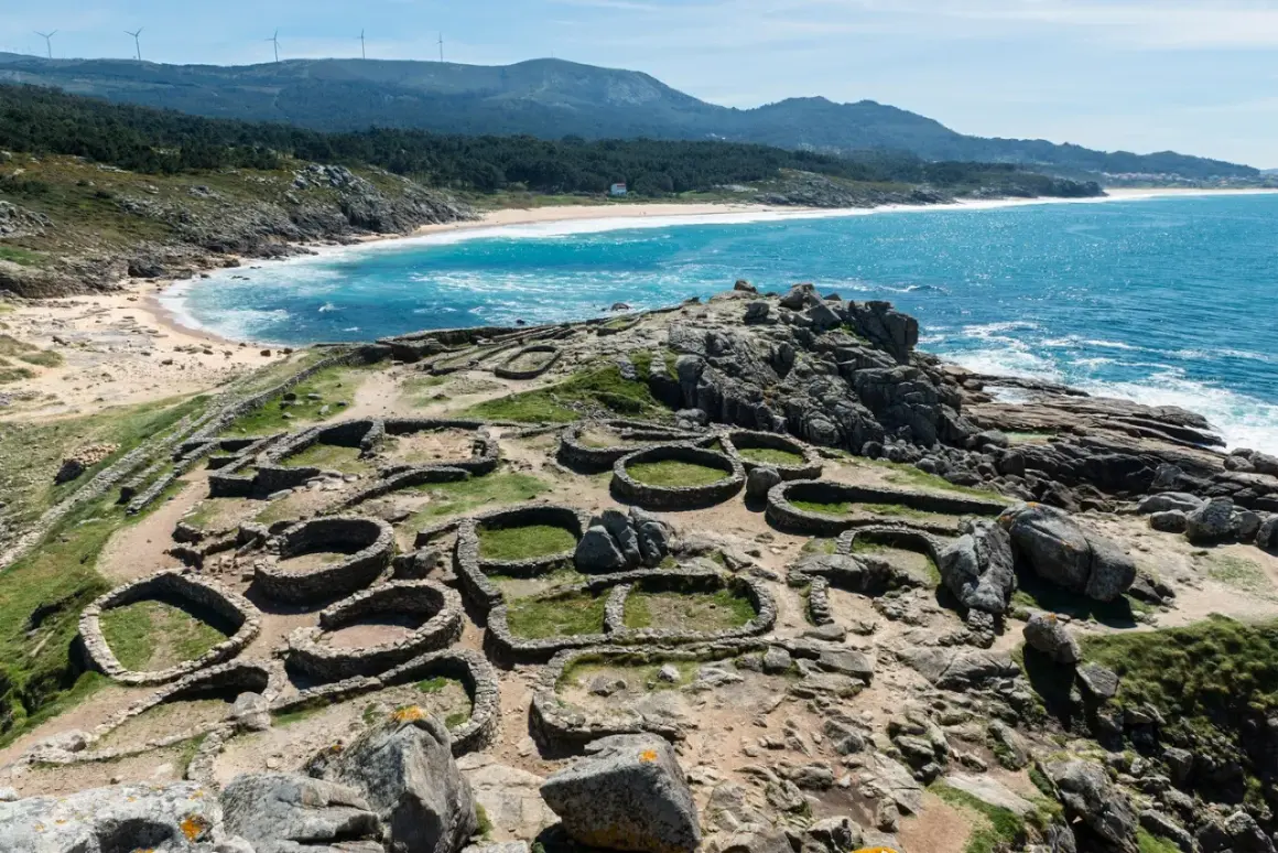 Ruinas circulares de piedra, vestigios de antiguos poblados, se extienden sobre un promontorio rocoso con el mar azul al fondo. Estos son algunos de los monumentos de Galicia.