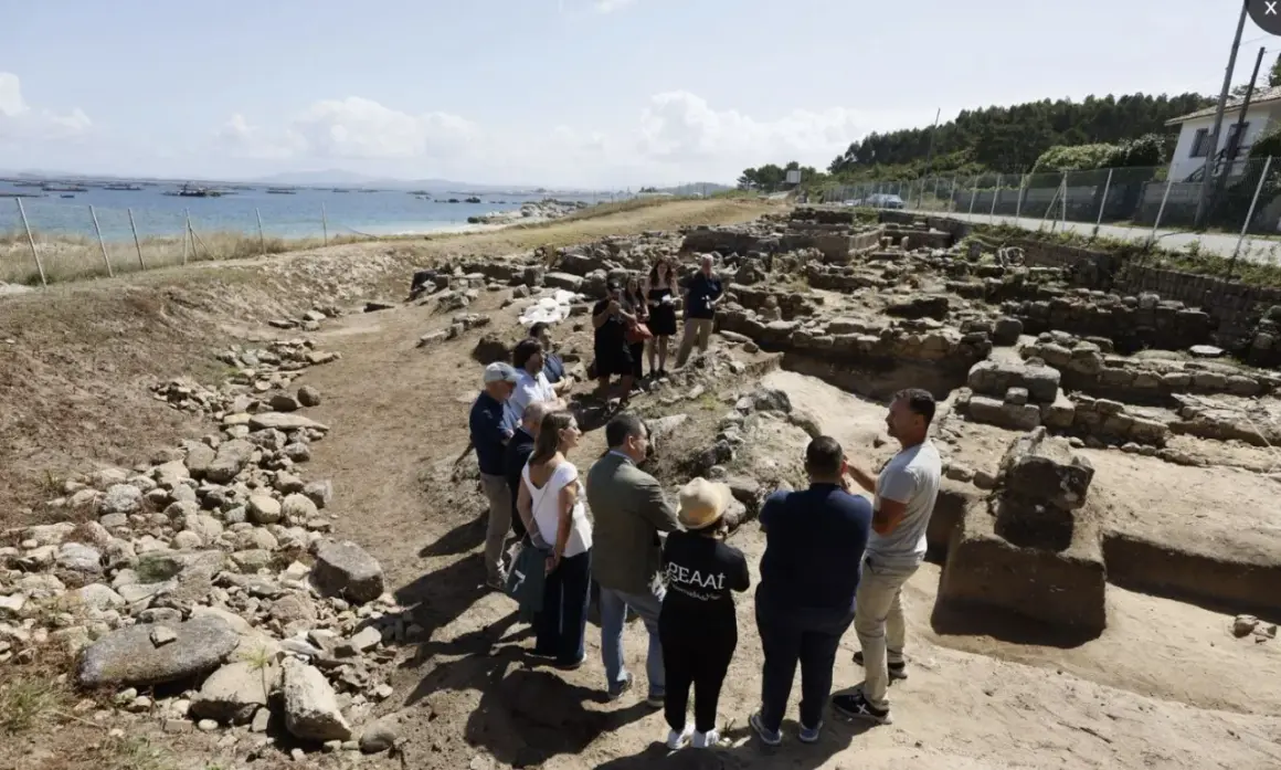 Grupo de personas observando ruinas arqueológicas en la costa. Se aprecia el mar y el **adro vello** del yacimiento.